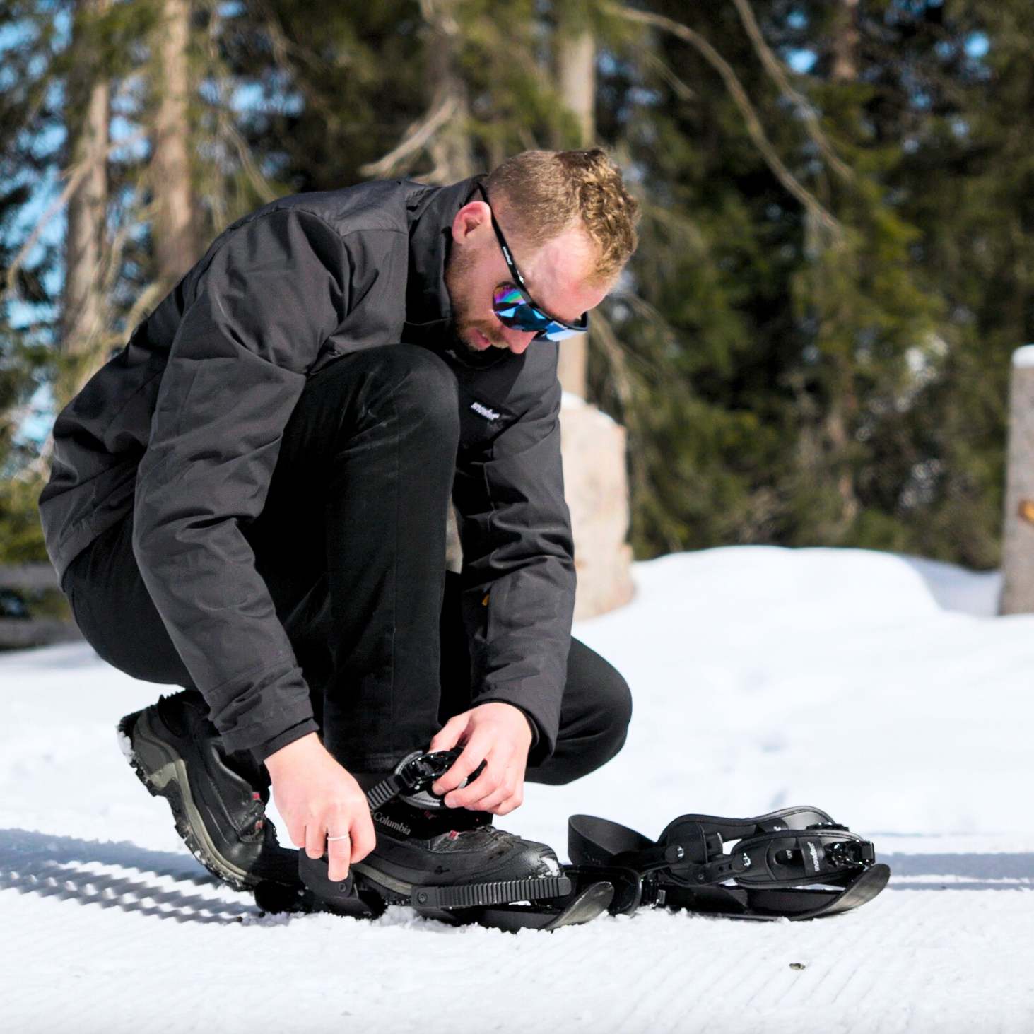 Person adjusting snowshoes in a snowy landscape with trees in the background