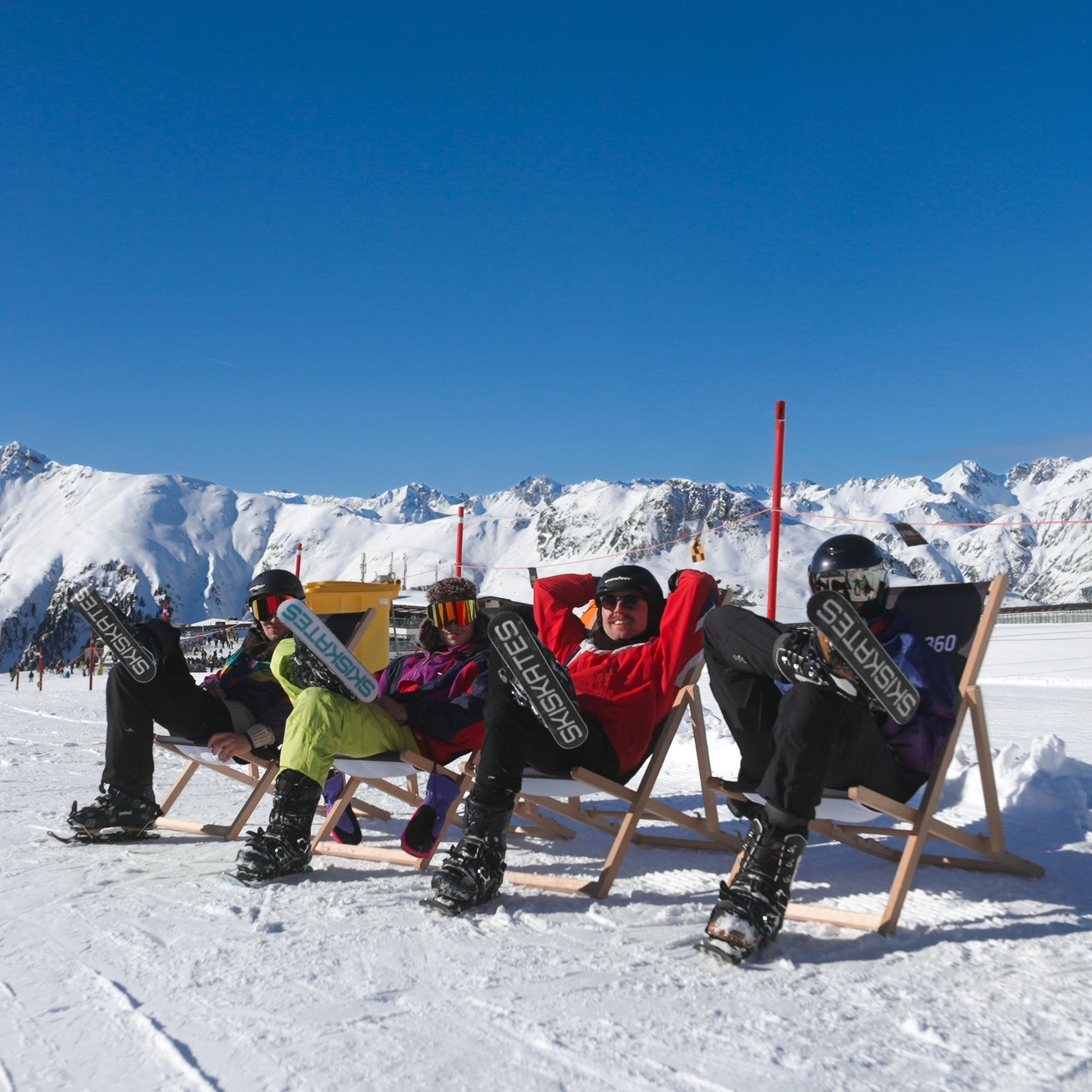 Four people sitting on chairs with snowboards, enjoying a break against a snowy mountain backdrop.