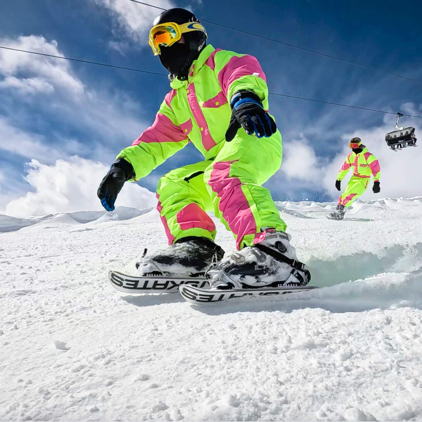 Person in bright green and pink snowsuit snowboarding on a snowy slope with another person in the background.